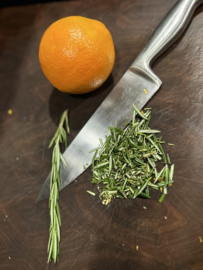 A whole orange and chopped fresh rosemary on a wooden cutting board with a knife to make the compound butter for smoked turkey.