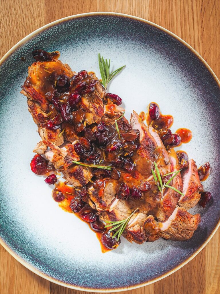 Cranberry Orange Pork Tenderloin sliced with orange cranberry sauce over it on a plate garnished with fresh rosemary.