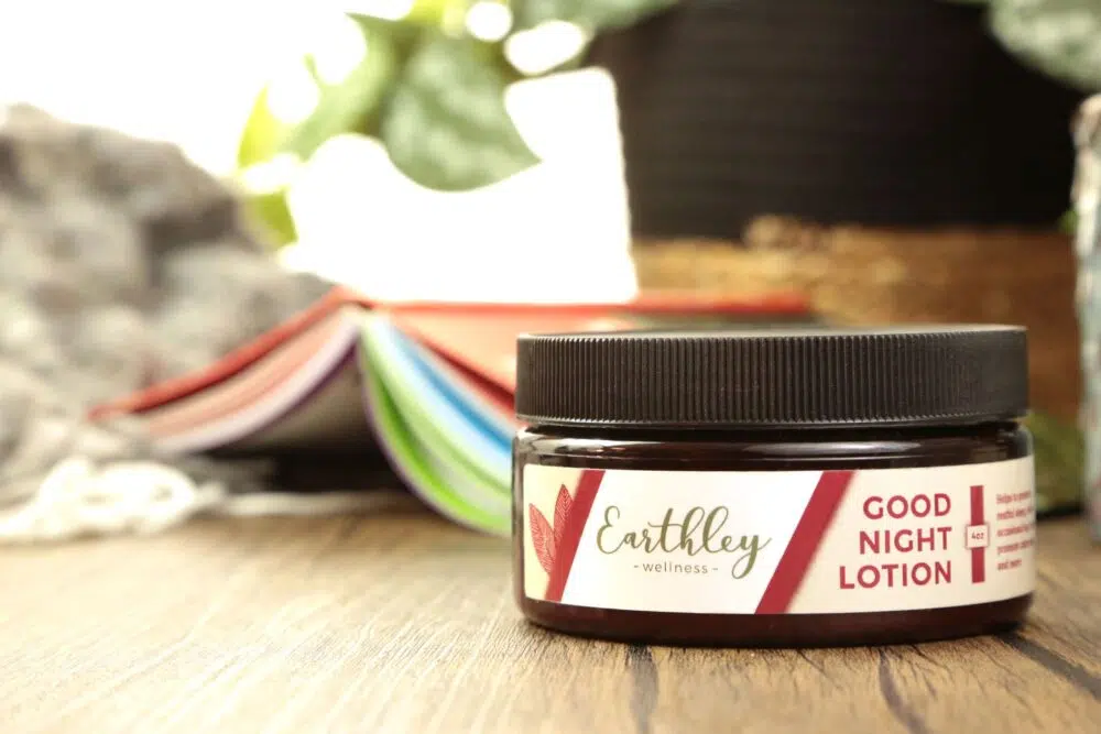 A jar of Good Night Lotion on a wooden counter with a book and plant in the background.