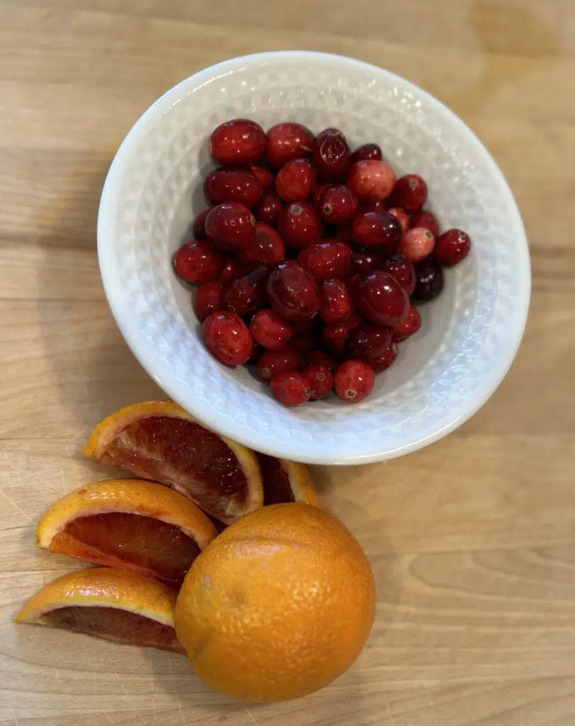 Fresh cranberries in a white bowl with a sliced blood orange on a wooden cutting board.