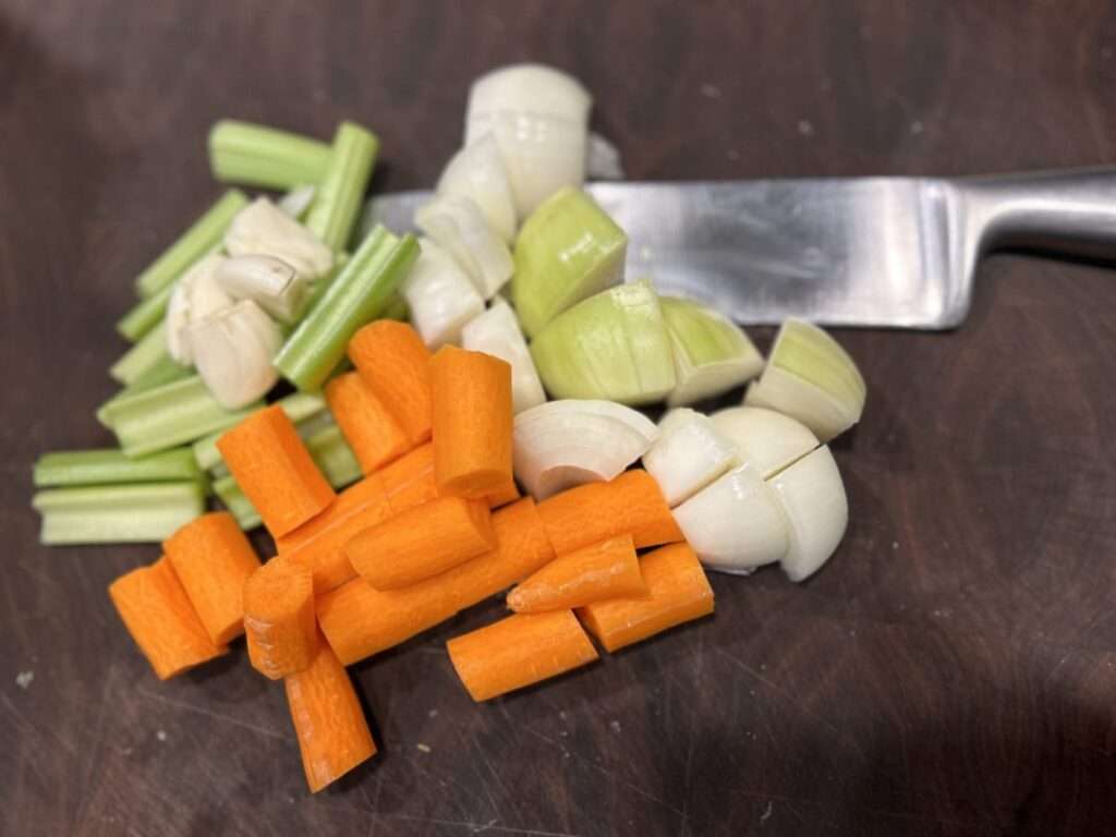 Chopped carrots, onions, garlic cloves and celery on a wooden cutting board with a knife.