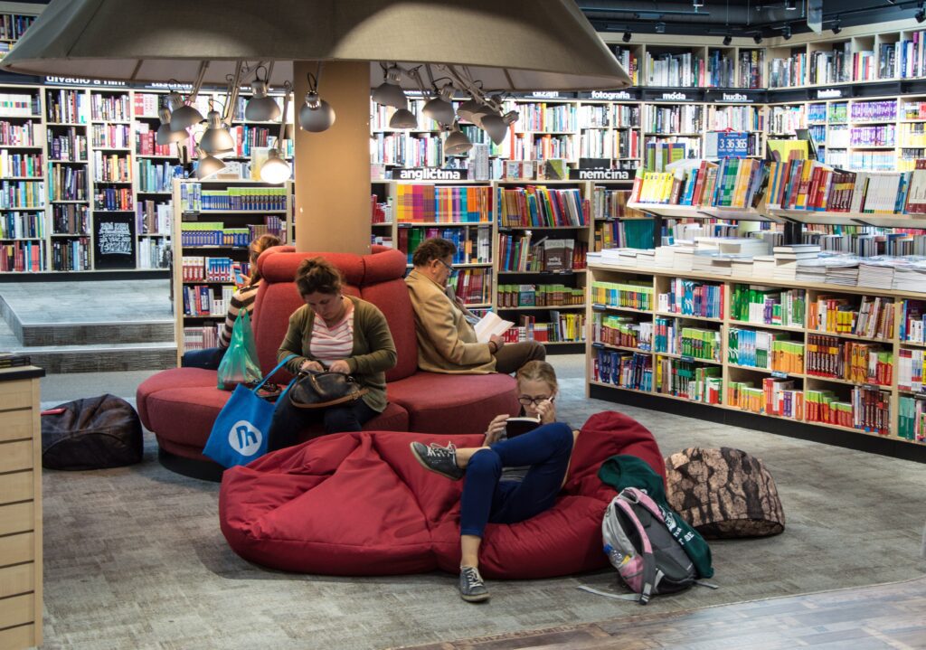 Kids reading books in a library on a red couch surrounded by backpacks.