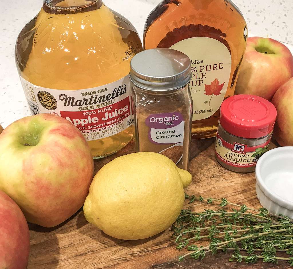 Apples, lemons, a jar of ground cinnamon and all spice and a bottle of real maple syrup with fresh thyme on a wooden cutting board.