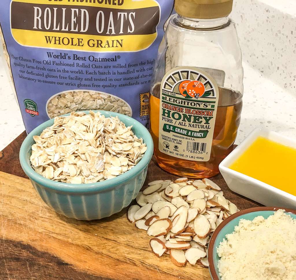A bag of whole grain rolled oats, honey, butter, and sliced almonds on a wooden cutting board.