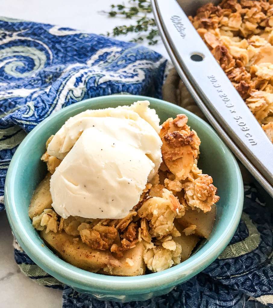A serving bowl of sugar free apple crisp topped with vanilla ice cream next to the pan of apple crisp fresh from the oven.