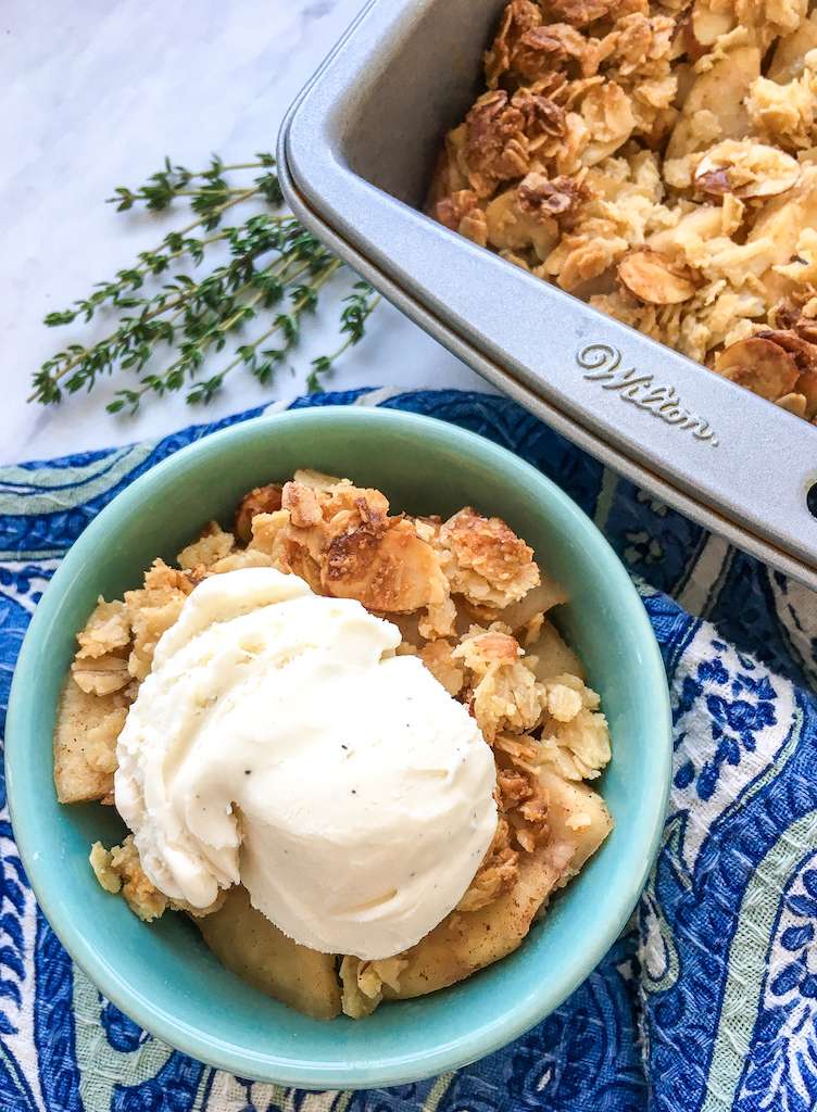 A serving of sugar free apple crisp in a bowl next to the pan of apple crisp with fresh thyme on the side.