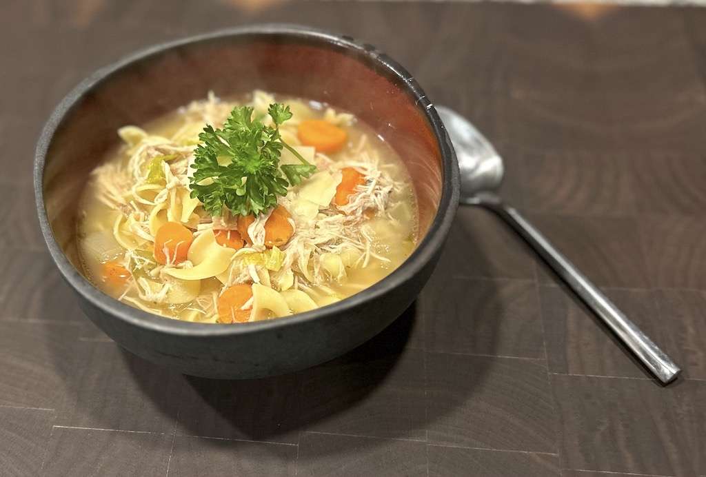 A bowl of my secret ingredient chicken noodle soup on a wooden cutting board with a spoon.