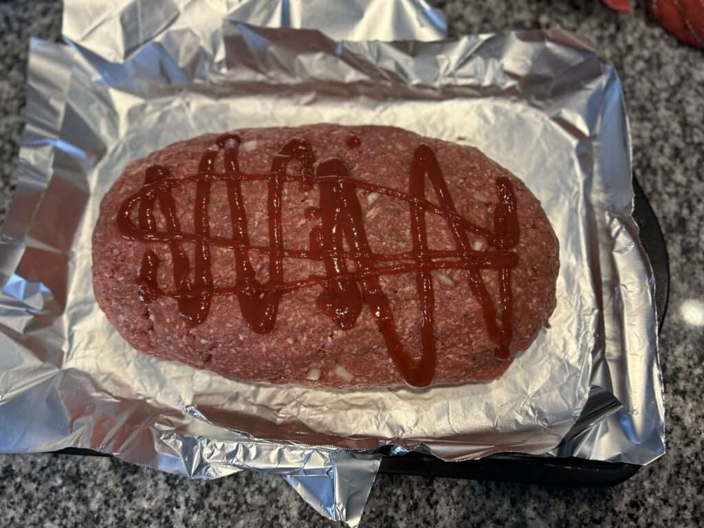A 6 pound meatloaf drizzled with ketchup on a tin foil lined cookie sheet ready to be smoked on the grill.