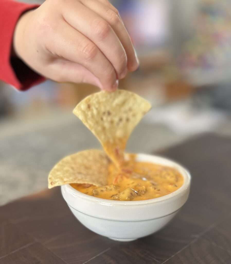 A picture of my sons hand dipping a tortilla chip into 3 ingredient queso with chorizo.