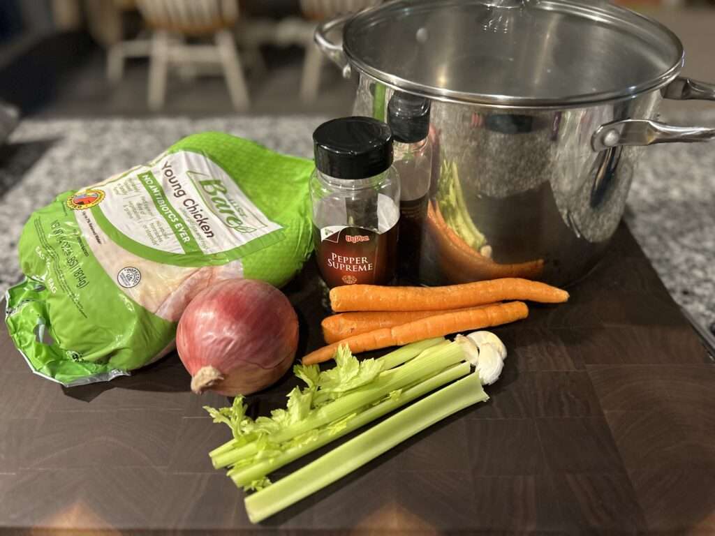 A wooden cutting board with a large stock pot surrounded by a whole chicken, carrots, celery, peppercorns and an onion.