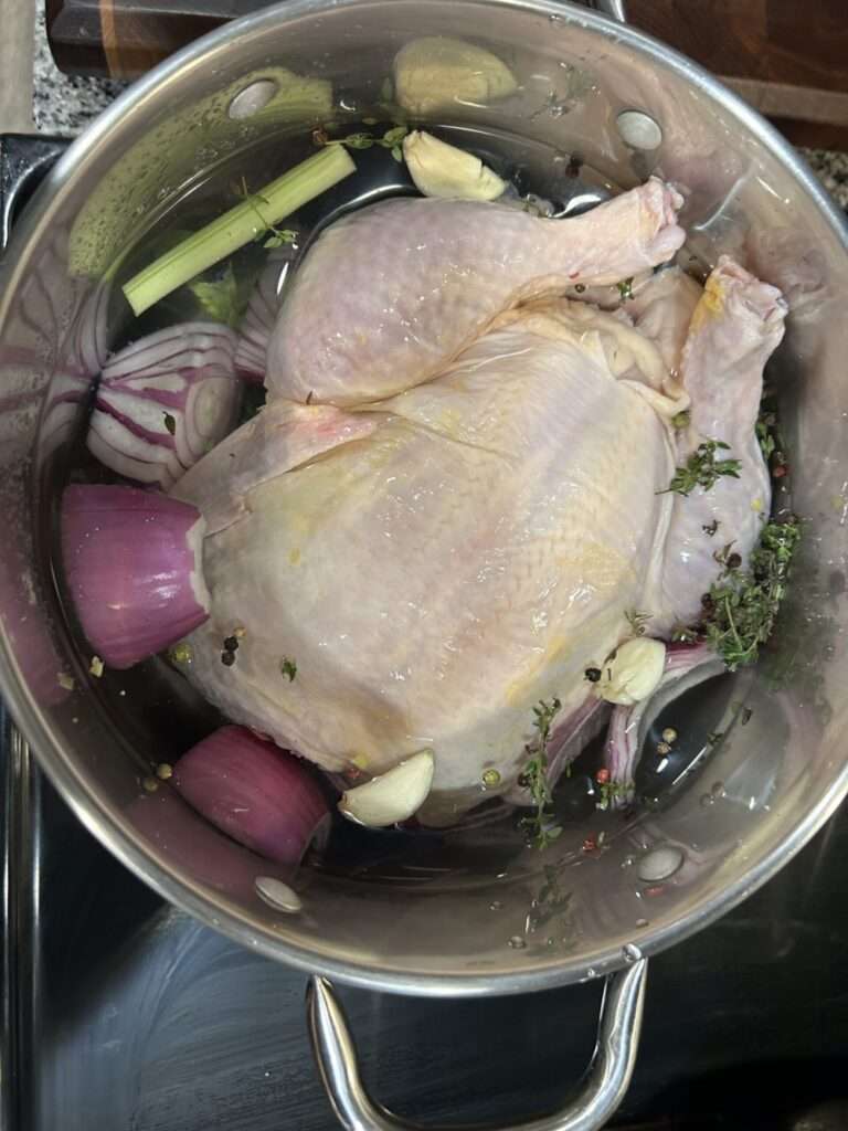 A whole chicken raw in a large stock pot filled with water, peppercorns, red onions, celery, fresh thyme and garlic cloves.