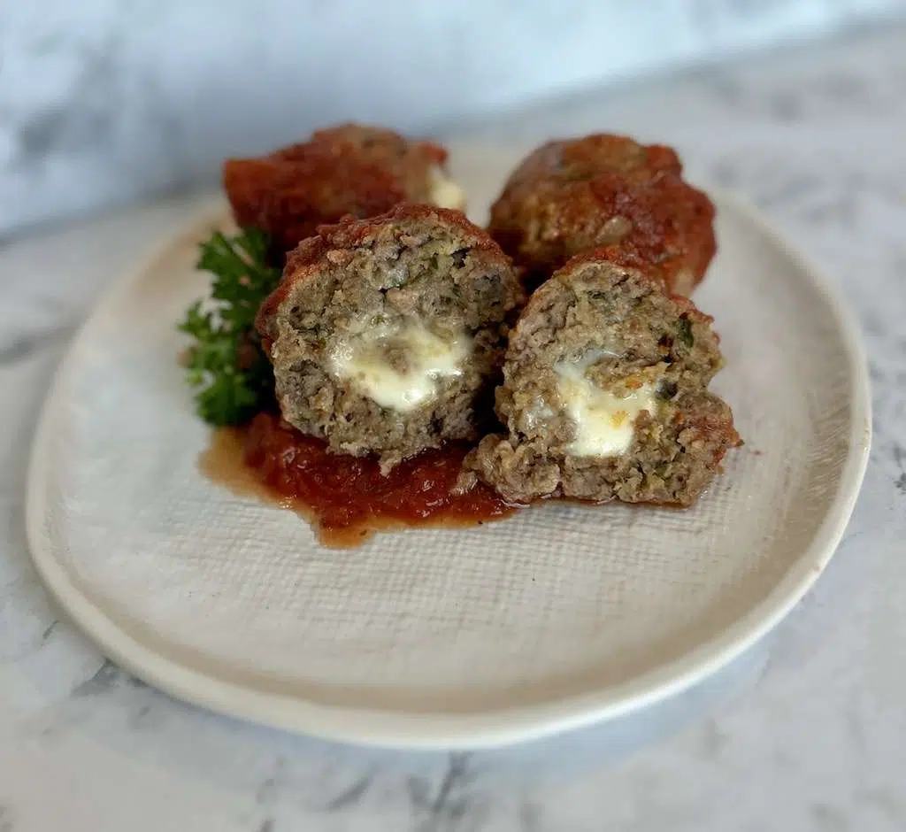 A white plate of mozzarella stuffed meatballs garnished with fresh parsley on a pool of marinara sauce.