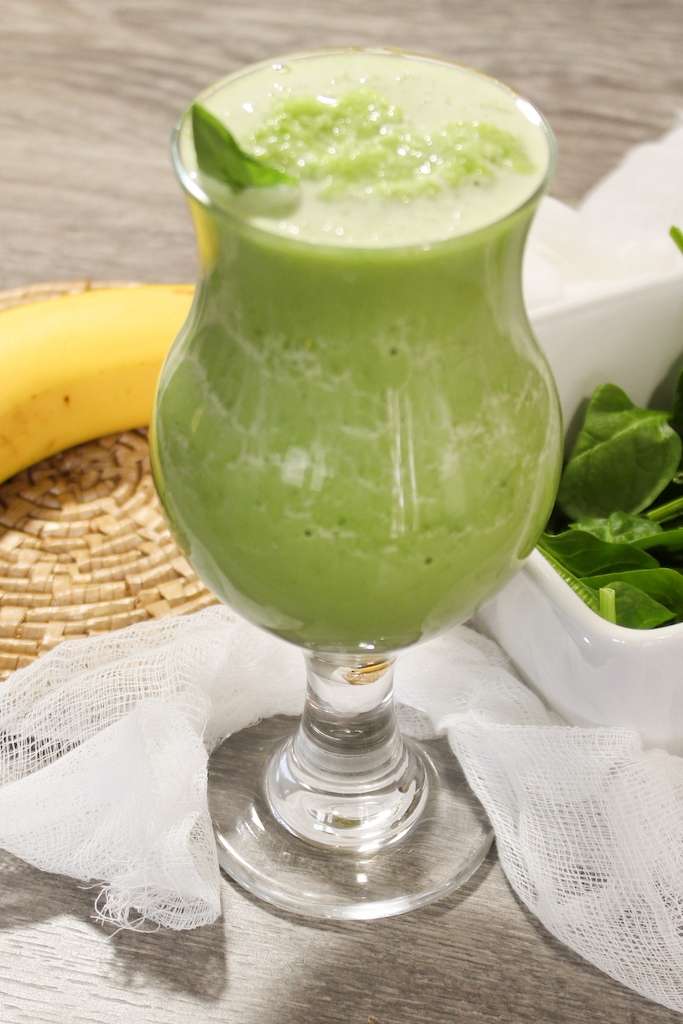 A tall glass of spinach banana smoothie with a banana and a bowl of fresh spinach leaves on the side.