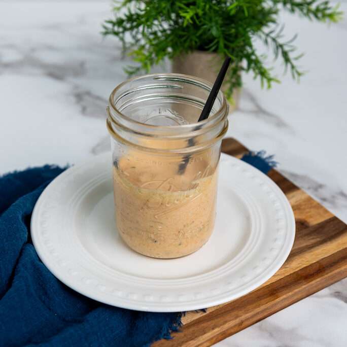 A mason jar of big mac sauce half filled with a black spoon for serving on a white plate with a fern in the background.