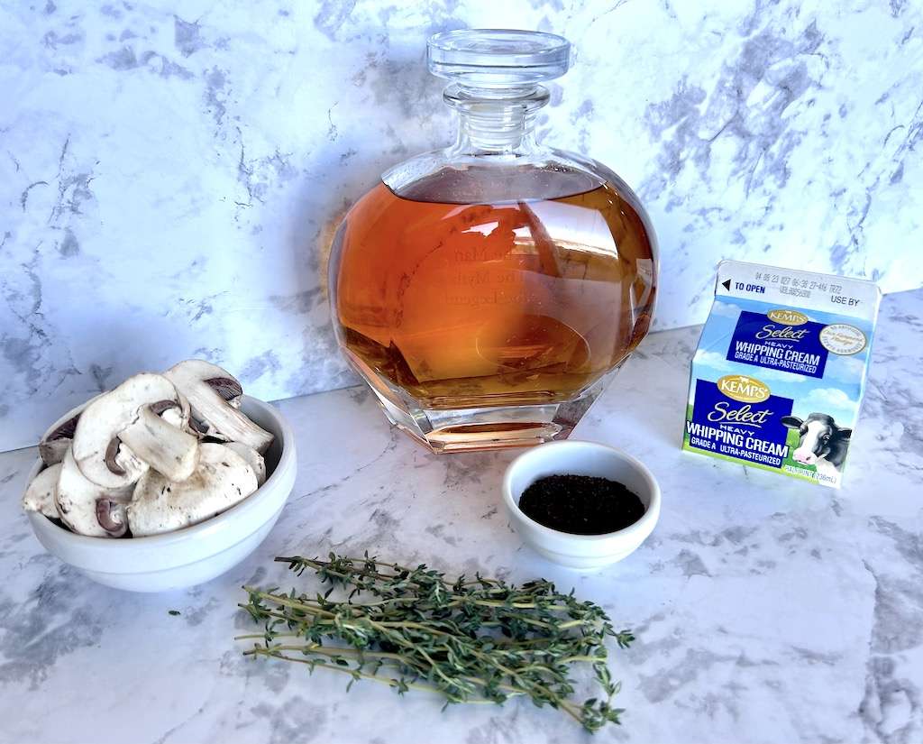Coffee Rubbed Tri-Tip Steak Ingredients on a marble counter top including a bourbon decanter, heavy whipping cream. fresh sliced mushrooms, ground coffee, and fresh thyme.