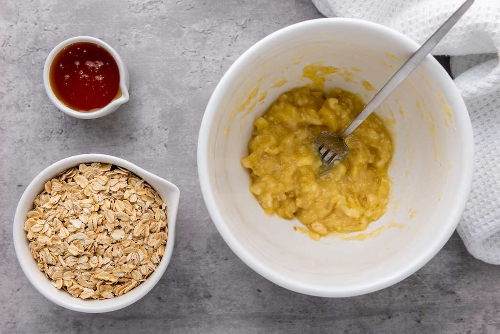 Oats and honey next to a mashed banana in a white mixing bowl.