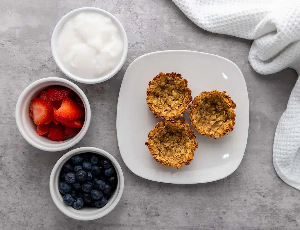 Granola Berry Cups - A Delicious Treat Mini granola cups on a white plate next to white bowls of yogurt, strawberries and blueberries.