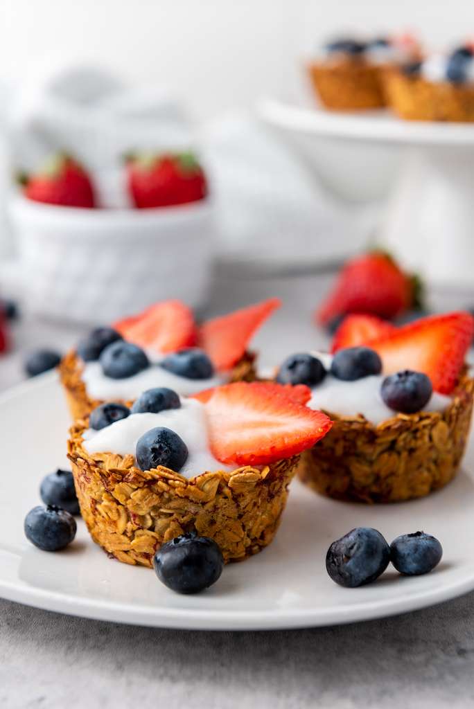 Three granola cups filled with vanilla yogurt topped with fresh sliced strawberries and blueberries on a white plate.