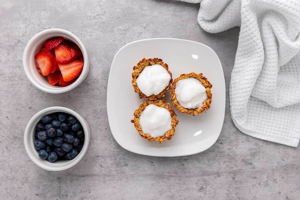 Mini granola cups filled with vanilla yogurt on a white plate next to small white bowls of sliced strawberries and blueberries.