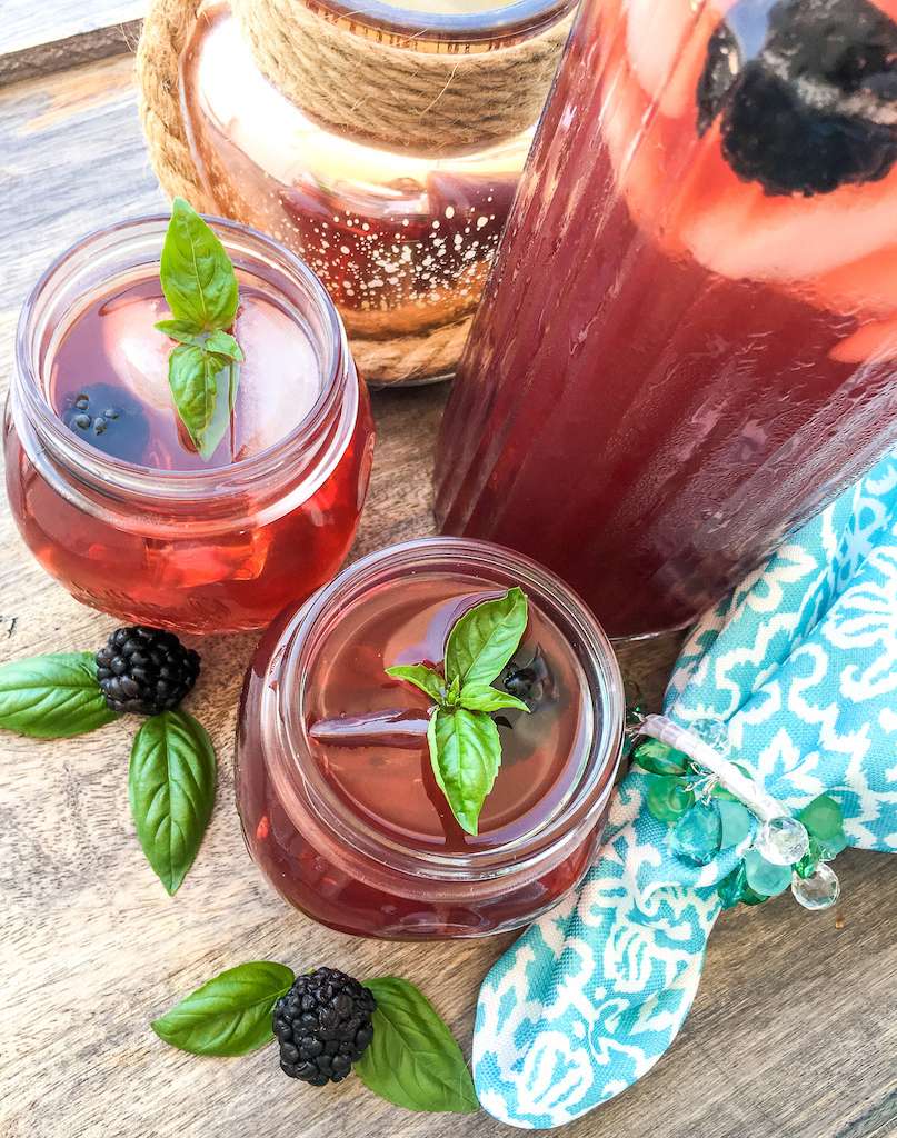A large pitcher of blackberry iced tea with two glasses of tea and fresh mint leaves and blackberries as a garnish.