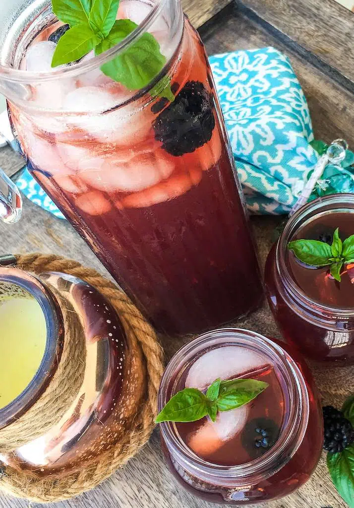 A large pitcher of blackberry iced tea with two small glasses of tea on a picnic table with a blue floral napkin.