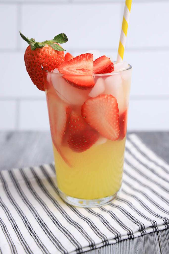 A tall glass of limoncello strawberry lemonade garnished with a fresh strawberry and a paper straw on a striped napkin.