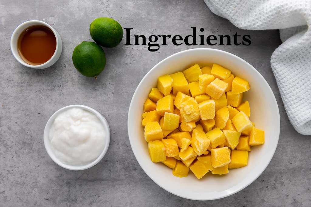 The ingredients for mango coconut lime popsicles including a bowl of frozen mango chunks, coconut yogurt, two fresh limes and a small bowl of agave syrup.