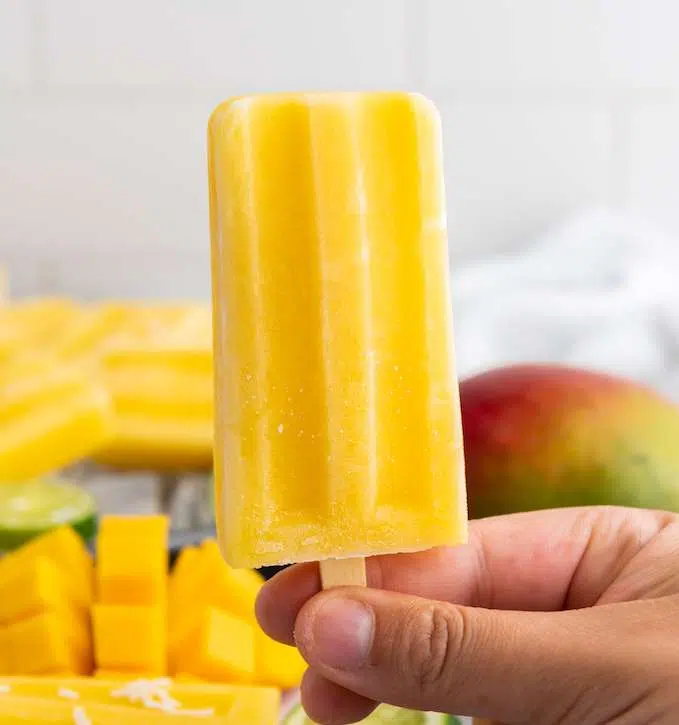Delicious Tropical Mango Coconut Lime Popsicle Recipe Mango Lime Popsicle on a stick with diced mangos in the background.