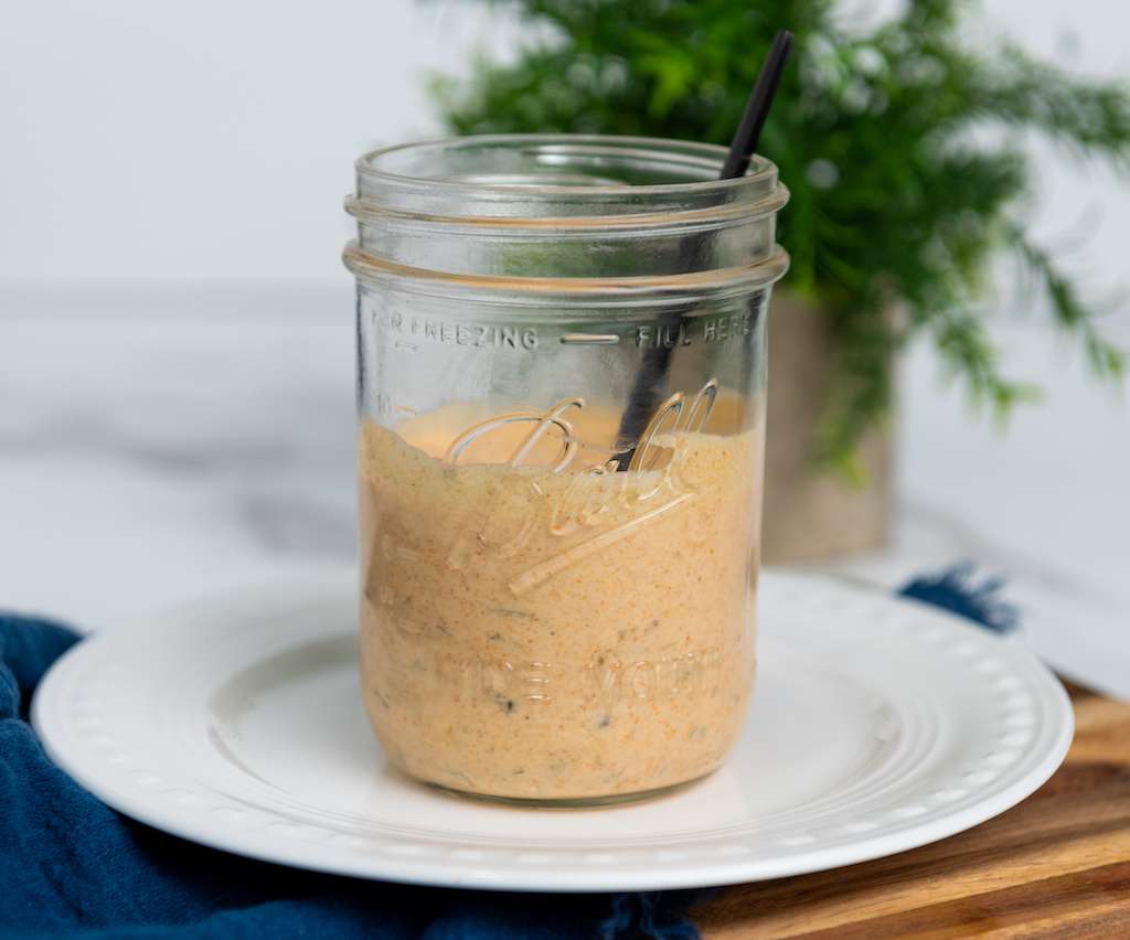 A glass mason jar half filled with big mac sauce with a spoon sticking out on a white plate with a fern in the background.