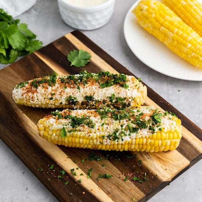 Two elotes or ears of easy Mexican street corn on a wooden cutting board.