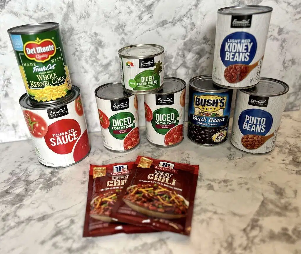 Cans of kidney beans, corn, diced tomatoes, tomato sauce, diced green chilis and two packets of chili seasoning on a marble counter top.