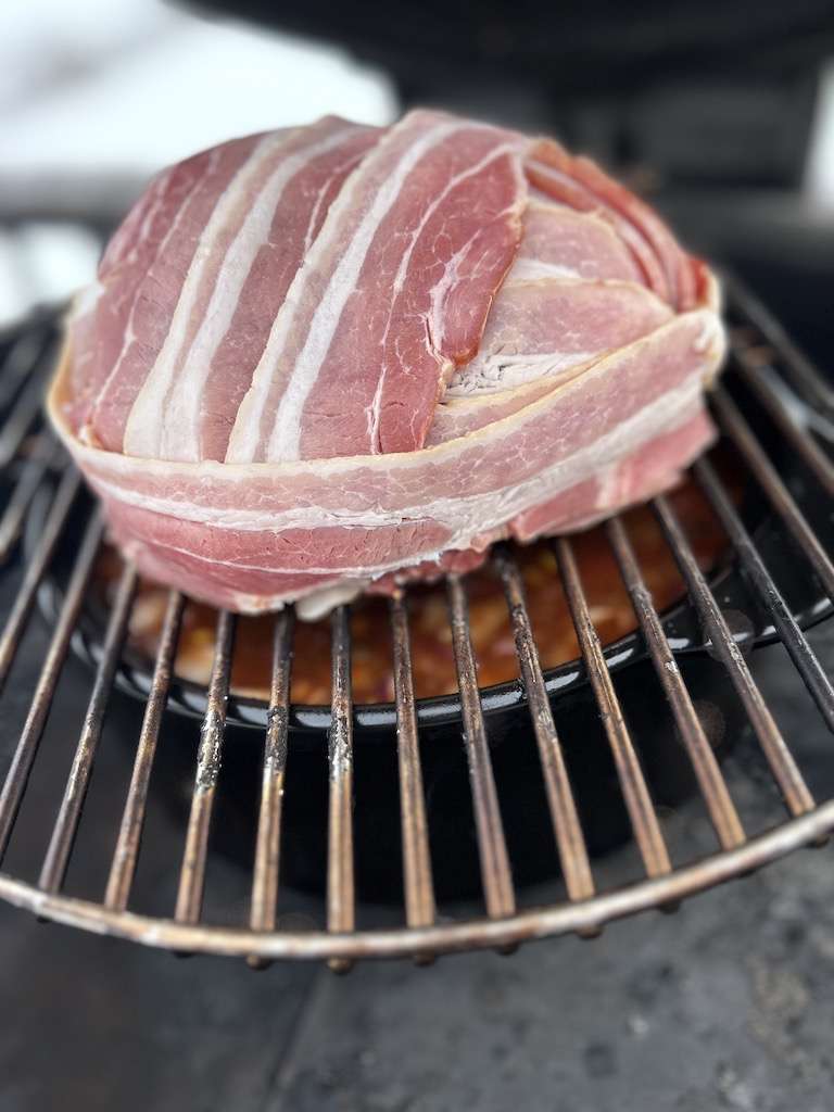 A large bacon wrapped meatball on a grate over the pot of over the top chili on the grill.