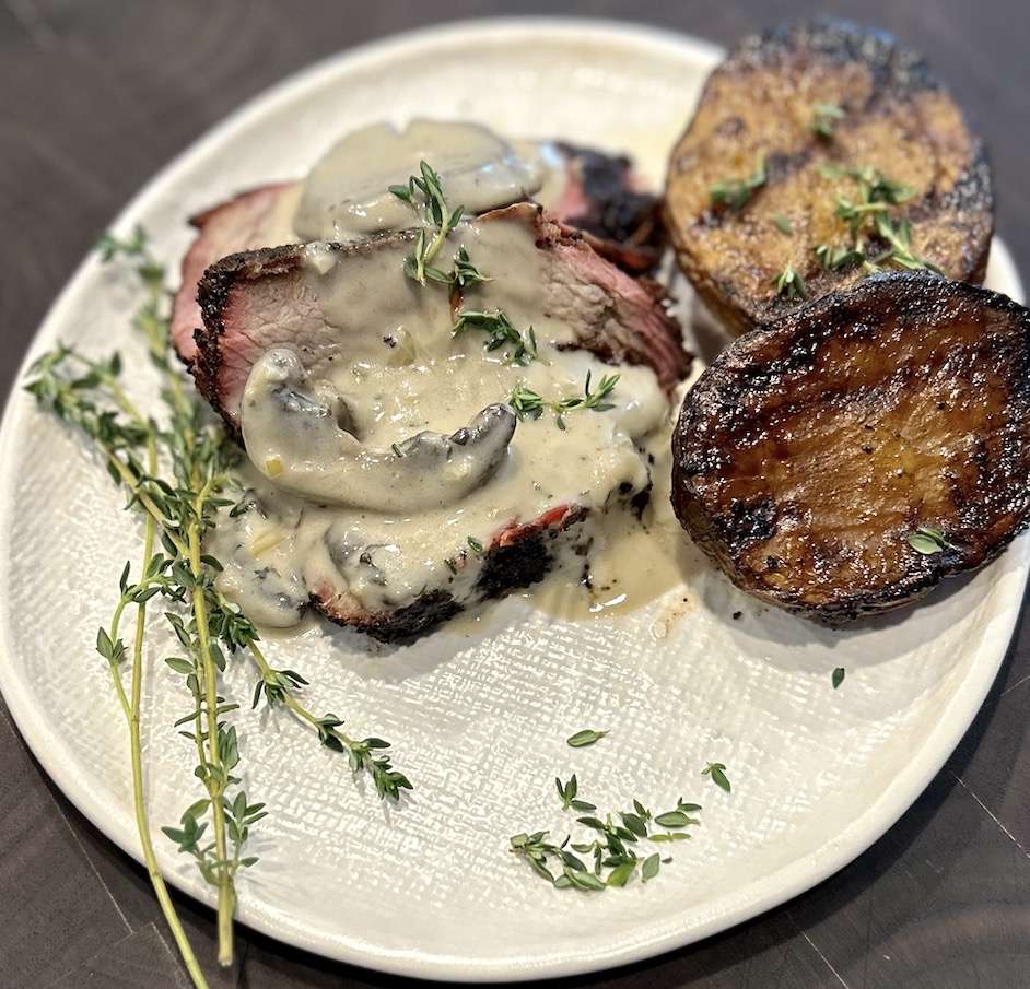 Plated Coffee Rubbed Tri-Tip drizzled with mushroom bourbon cream sauce with Grilled Potatoes on a white plate garnished with fresh thyme.