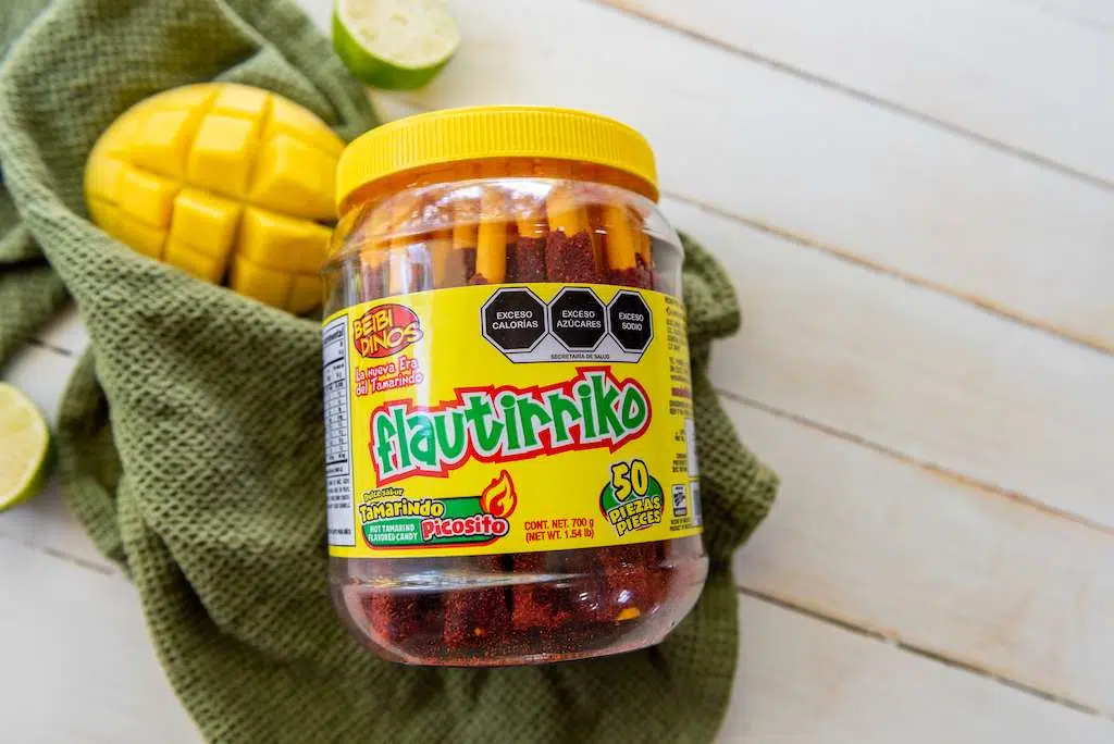 Tamarind Candy Sticks in a jar to use as garnish for chamoyada  with a sliced mango and a green towel on a wooden background.