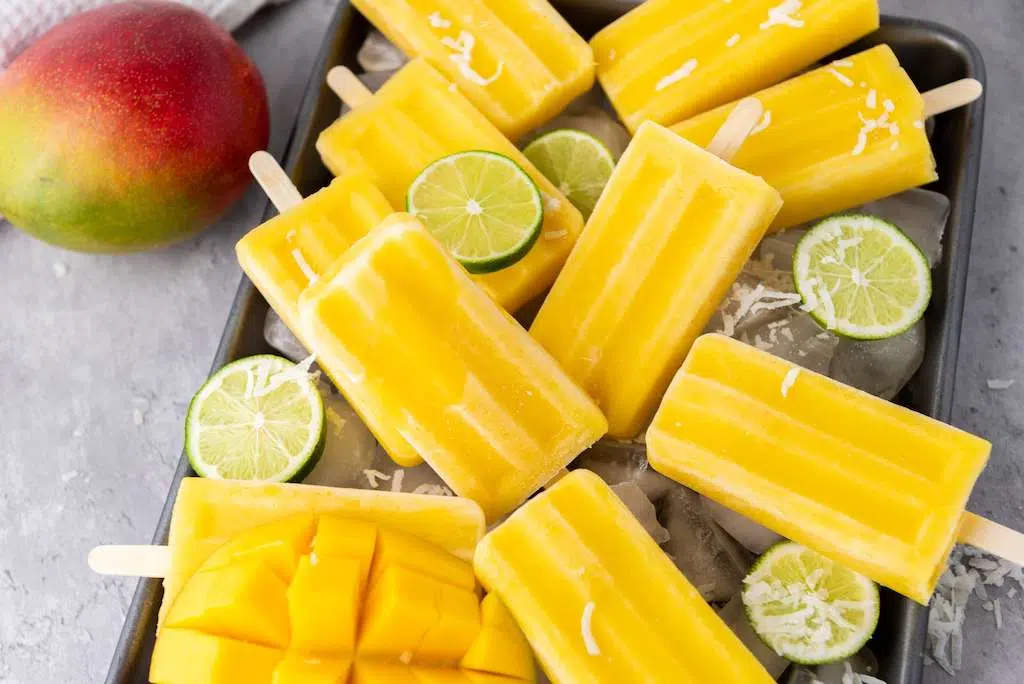 Delicious Tropical Mango Coconut Lime Popsicle Recipe Whole Mango with a cookie sheet of mango lime popsicles with fresh lime slices for garnish.