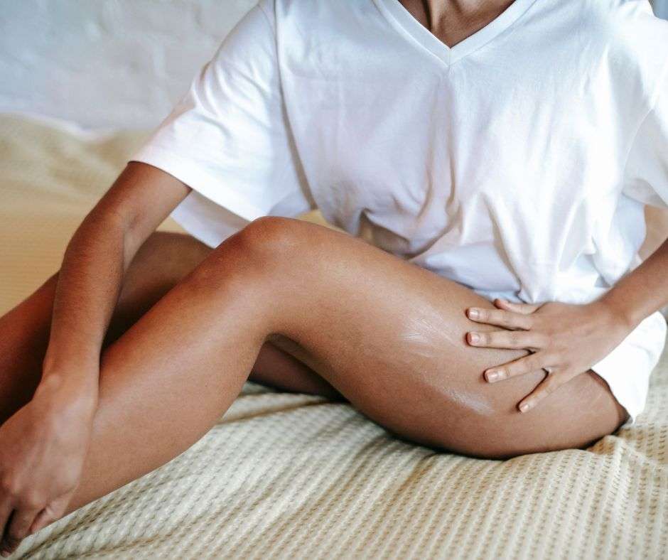 A women in a white t-shirt sitting on a bed rubbing her legs.