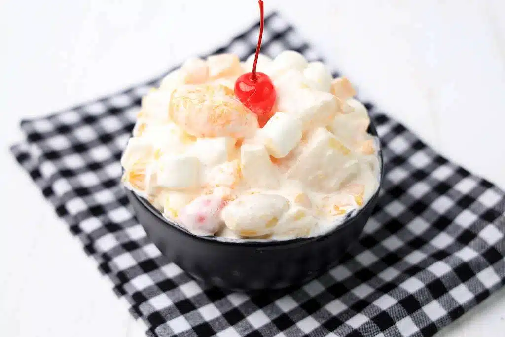 Ambrosia Salad with cool whip in a black bowl garnished with a cherry