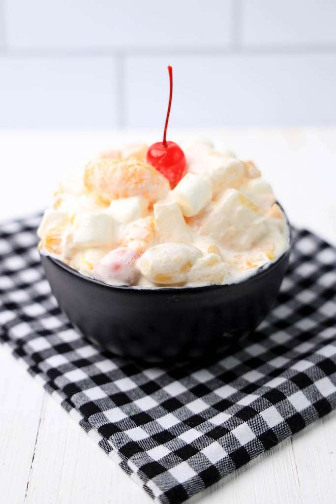 Ambrosia Salad with cool whip in a black bowl garnished with a cherry
