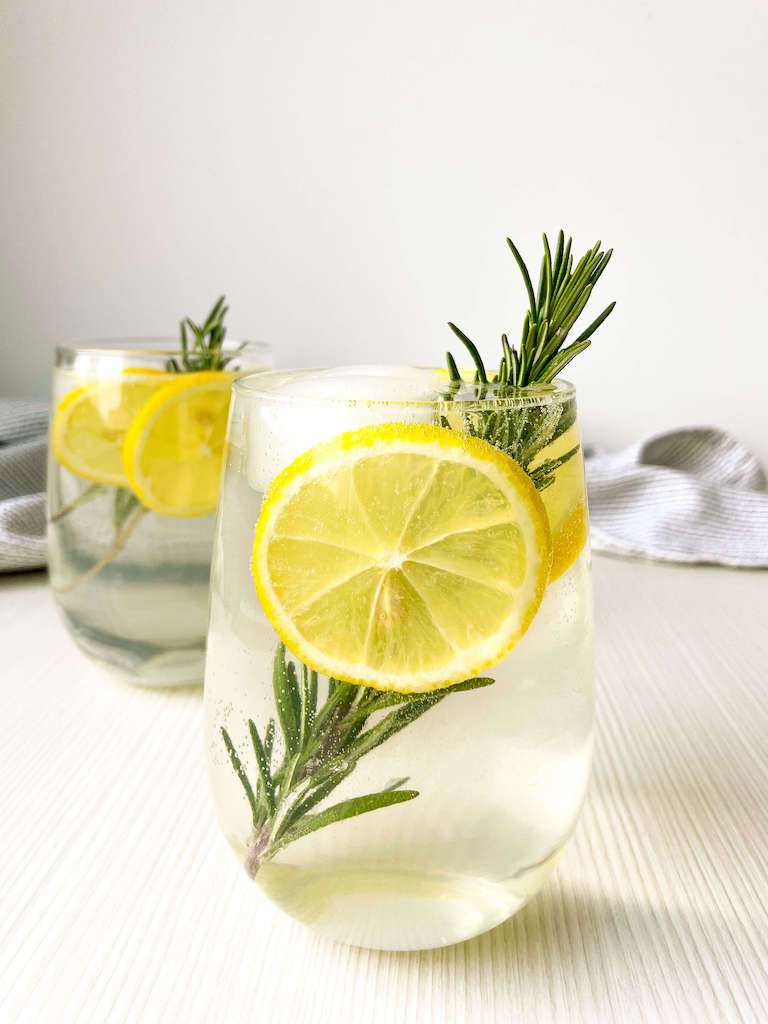 A glass of apple rosemary gin fizz with fresh lemon and rosemary with a 2nd glass in the background.