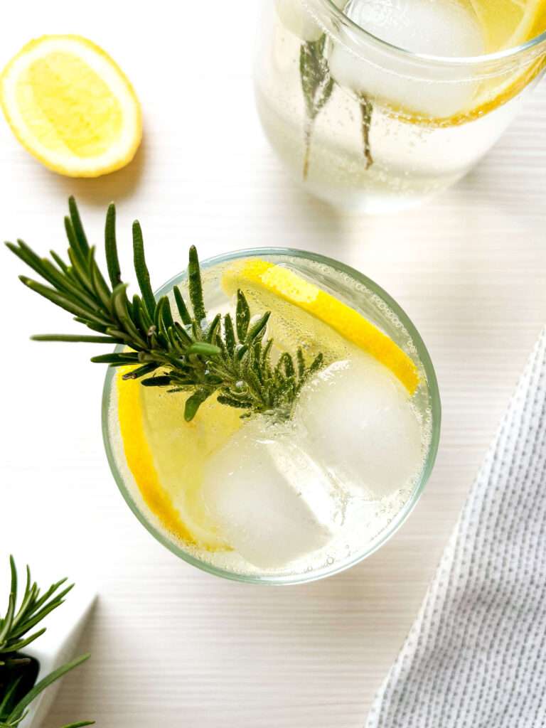 Looking down into a glass of apple gin fizz with fresh rosemary, lemon slices and ice.