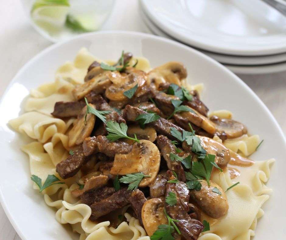 A white plate with egg noodles, a side dish for beef stroganoff, topped with beef stroganoff.
