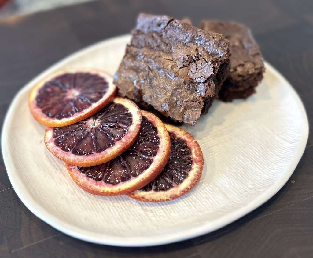 Delicious fresh blood orange slices on a white texture plate sitting on a wooden cutting board with two blood orange brownies.