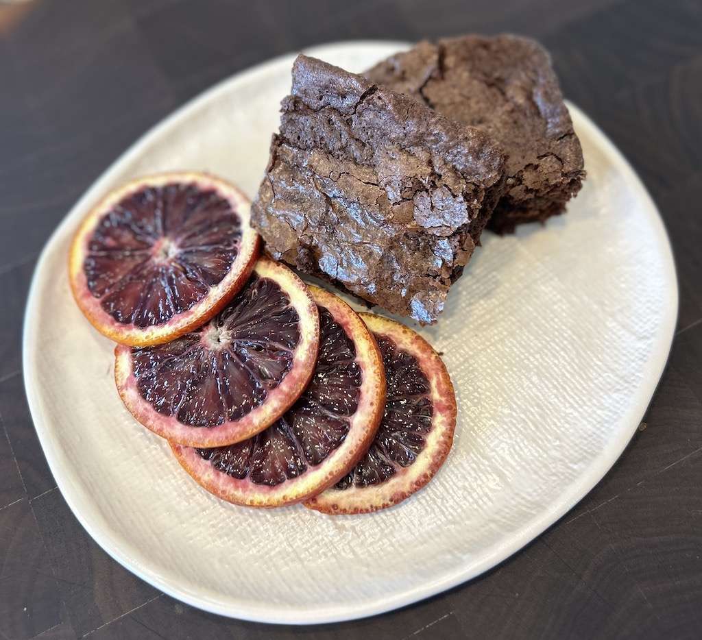 Two blood orange brownies on a textured plate with slices of fresh blood orange next to them as a garnish.