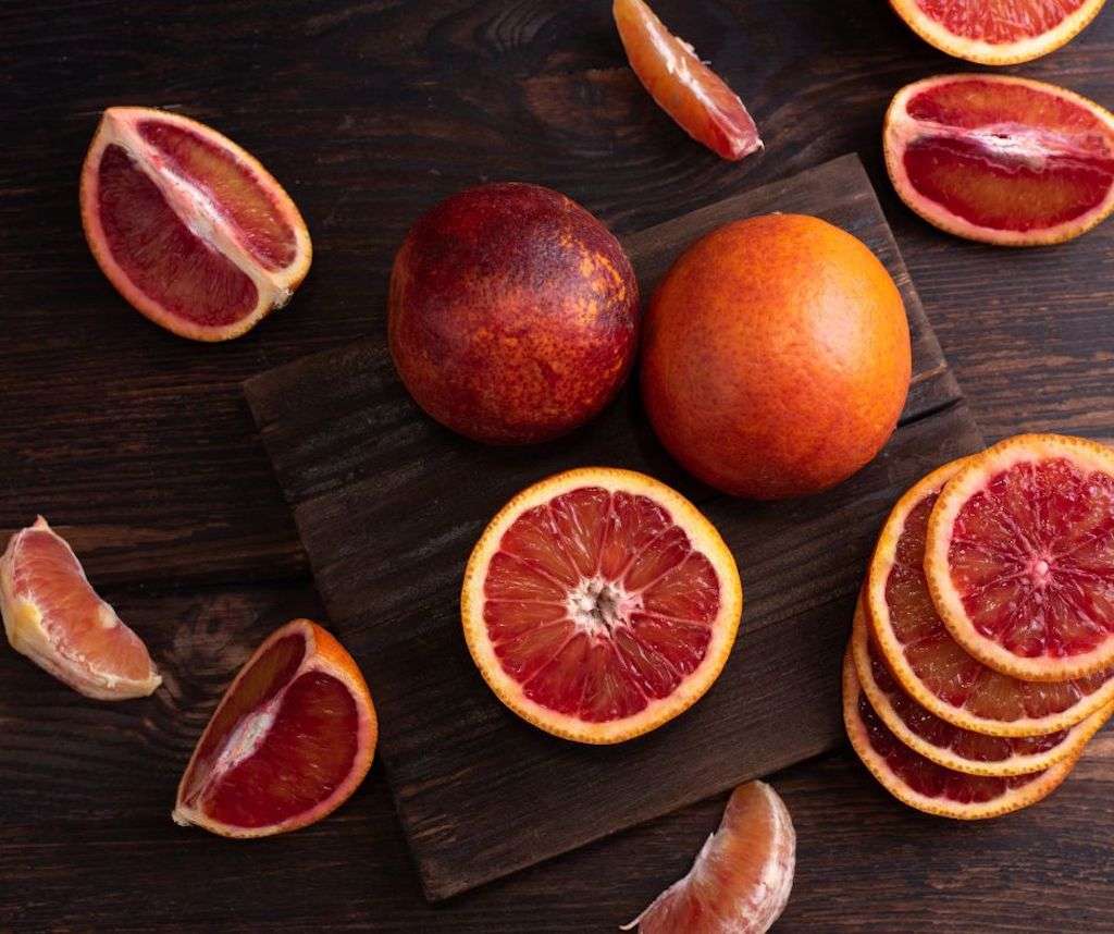 Two whole blood oranges and a few cut up blood oranges on a wooden cutting board.