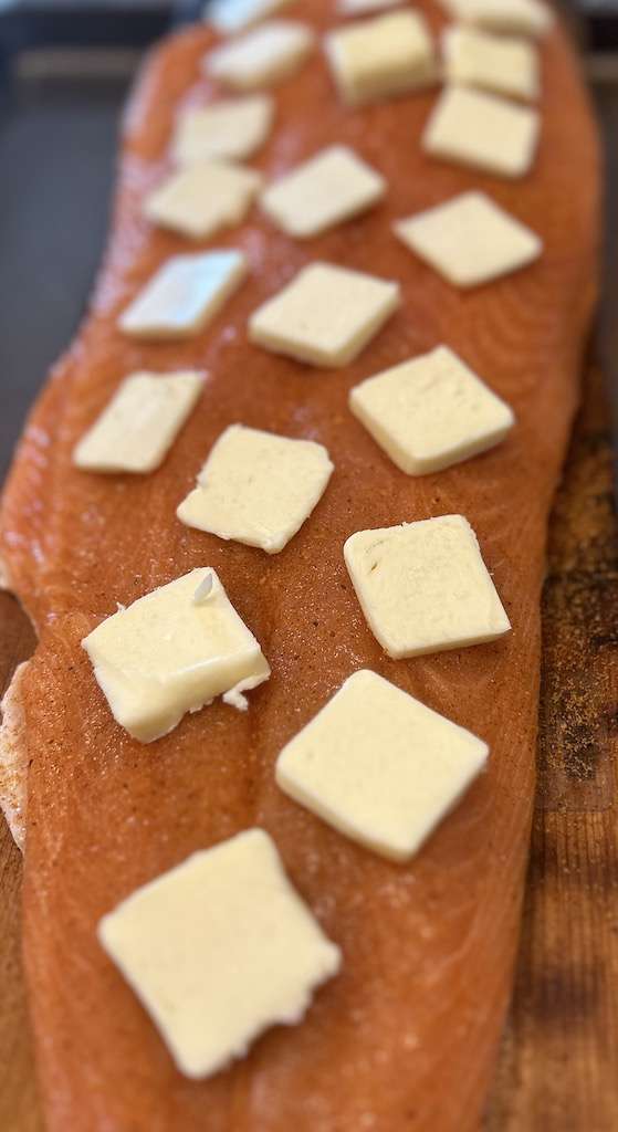 A seasoned salmon fillet topped with butter slices on a cedar plank.