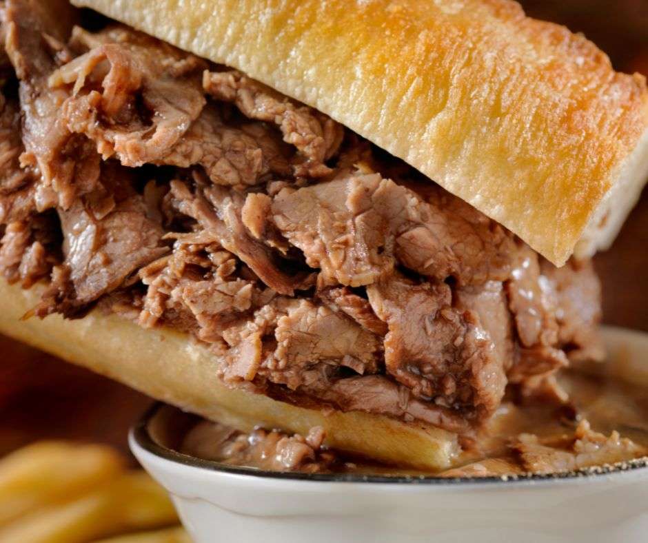 A french dip sandwich being dipped into au jus with french fries on the side for what to serve with french dip sandwiches.