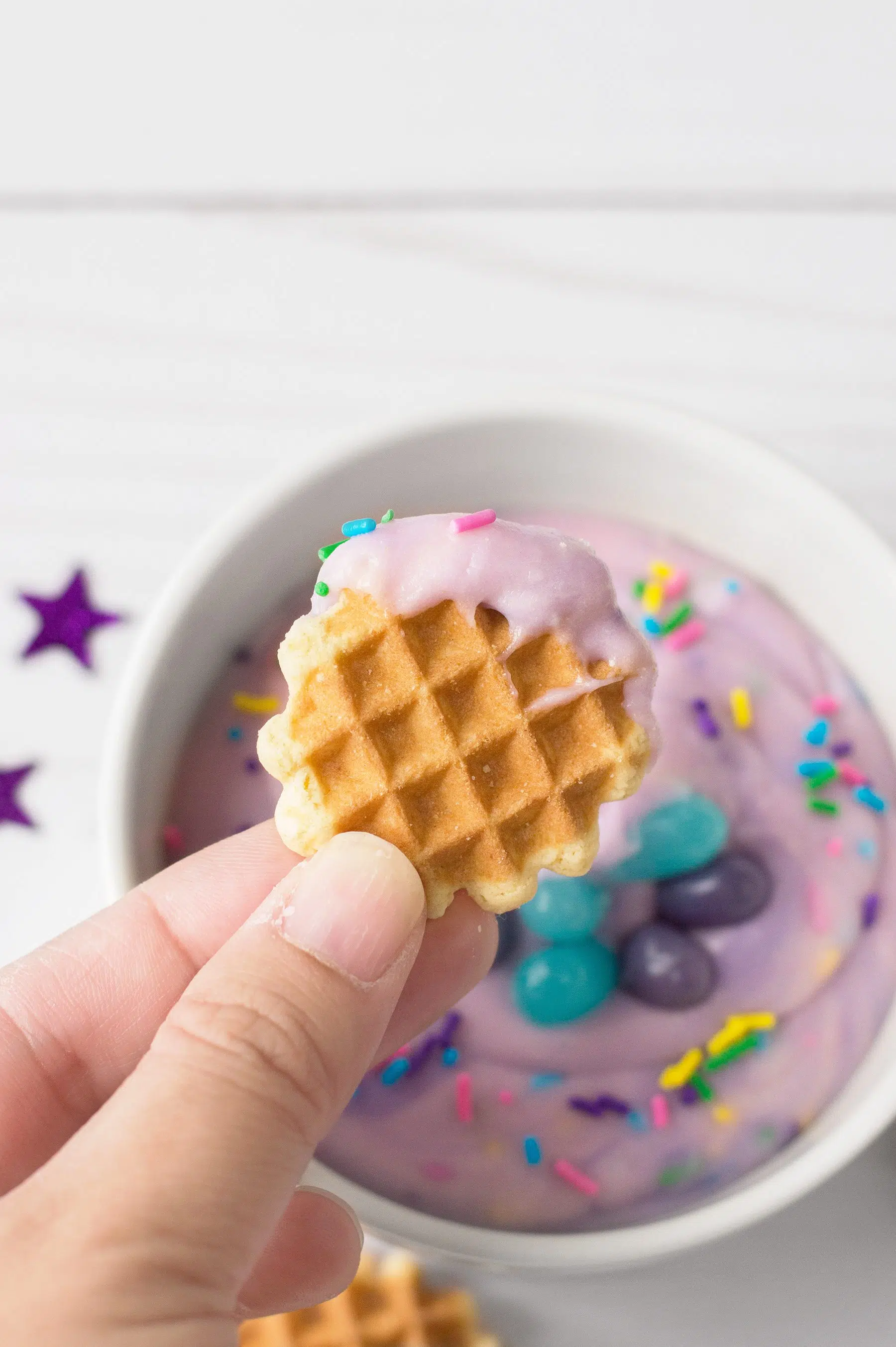Hand holding mini waffle cookie dipped in purple frosting with sprinkles