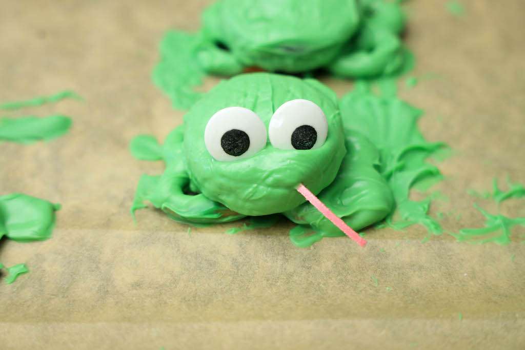 A green Oreo frog cookie assembled on parchment paper adding the candy eyes and tongue.
