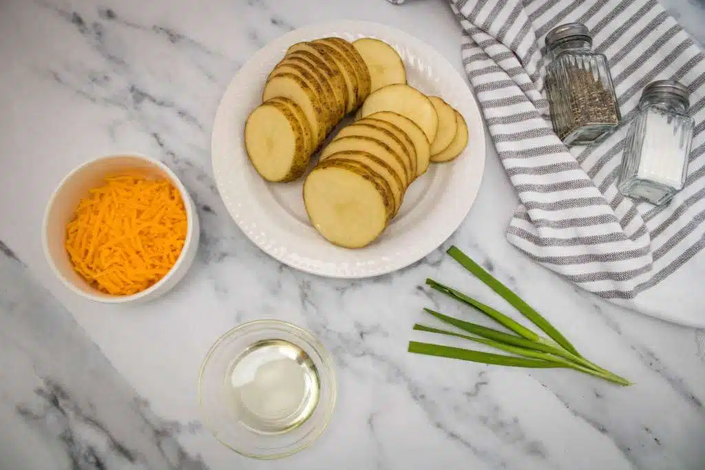 Sliced potatoes on a white plate with a bowl of cheddar cheese, a small glass of olive oil, salt and pepper shaker and fresh chives.