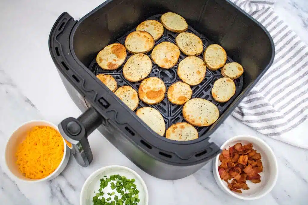 The air fryer with the potato slices lightly browned and a bowl of shredded cheddar cheese, crispy bacon and chopped fresh chives.